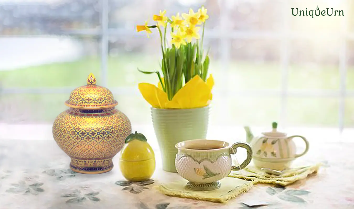 An elegant hand-painted UniqueUrn Benjarong art porcelain cremation urn with authentic 18k gold linework. The adult urn sits gracefully on a dining table beside a vase of yellow flowers, a teapot, and a cup set. Warm morning sunlight streams softly through sheer glass windows, emphasizing a serene, grounding place of honor within a luxury home.