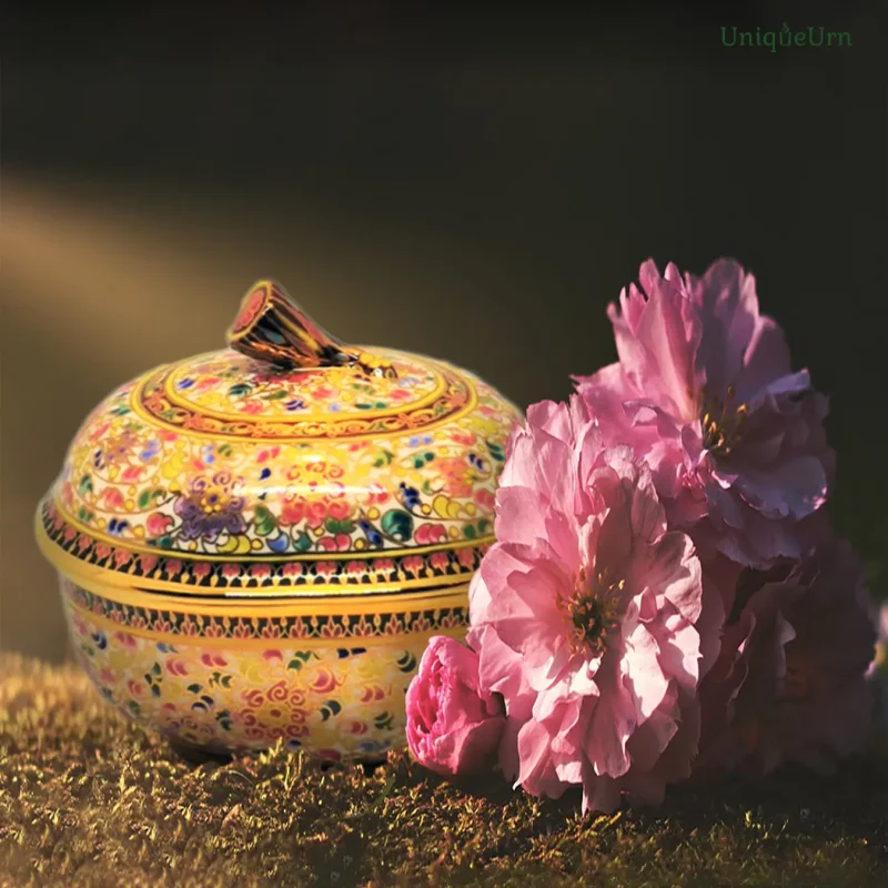 An elegant hand-painted UniqueUrn Bencharong art keepsake urn for ashes with authentic 18k gold linework. The oval keepsake urn sits gracefully on the grass beside pink flowers. Warm night light streams softly, emphasizing a serene, grounding place of honor.