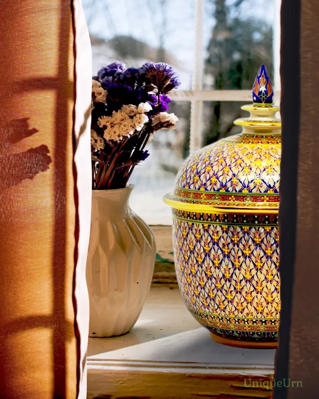 A master artisan's hand-painted geometry Phumkaobin Blue design on a porcelain Bencharong Thochan cremation urn sits on the window with sunlight.