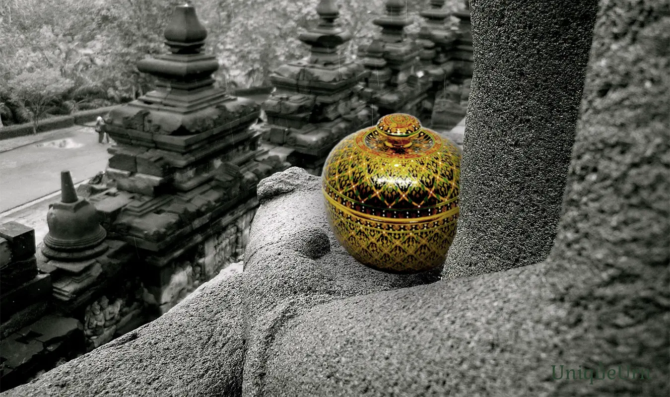 A master artisan's hand-painted geometry on a porcelain Bencharong keepsake box urn for ashes sits on the hands of a stone Buddha image.